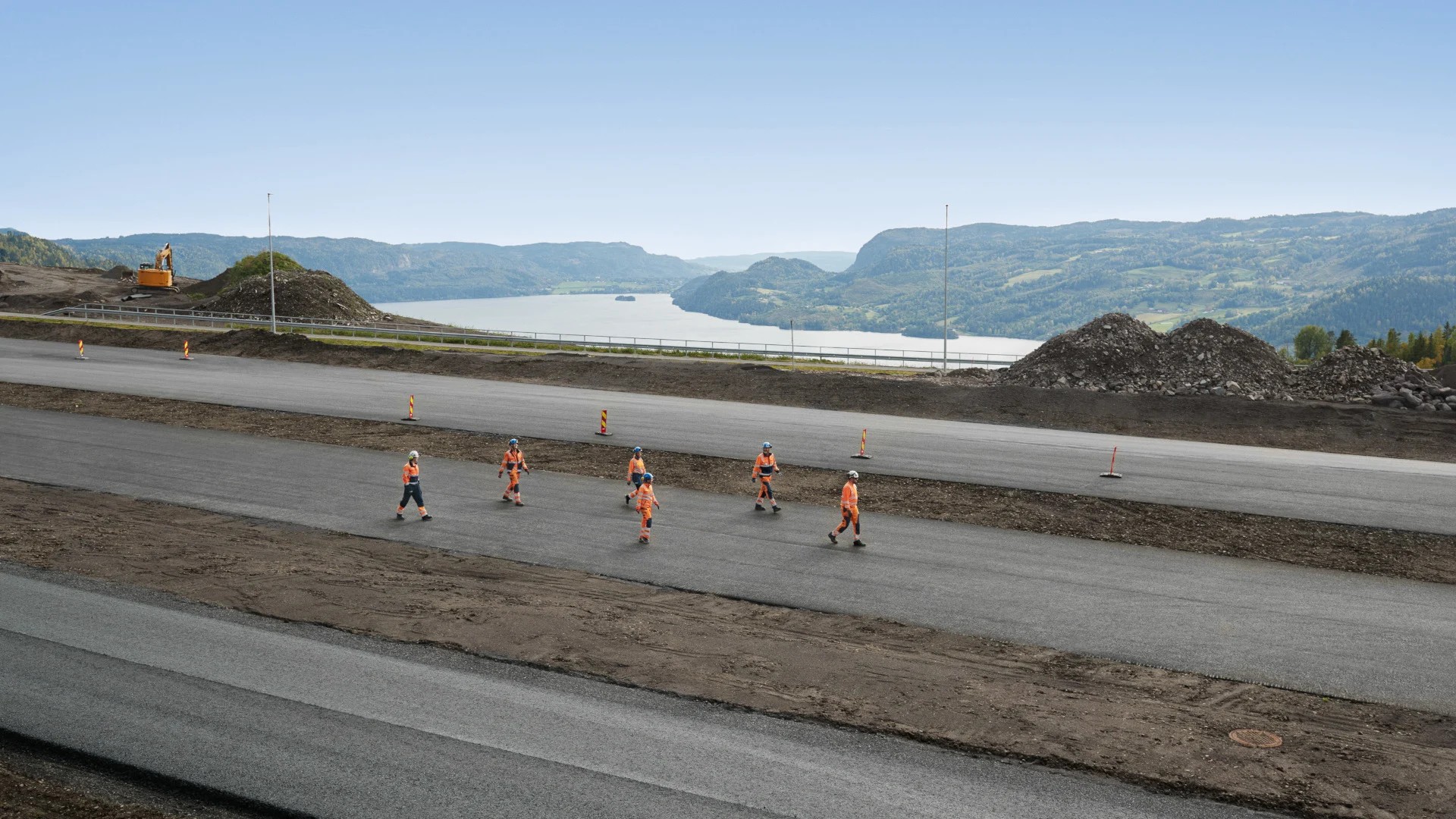 Byggnadsarbetare i orange säkerhetskläder som går på en stor asfalterad väg nära berg.