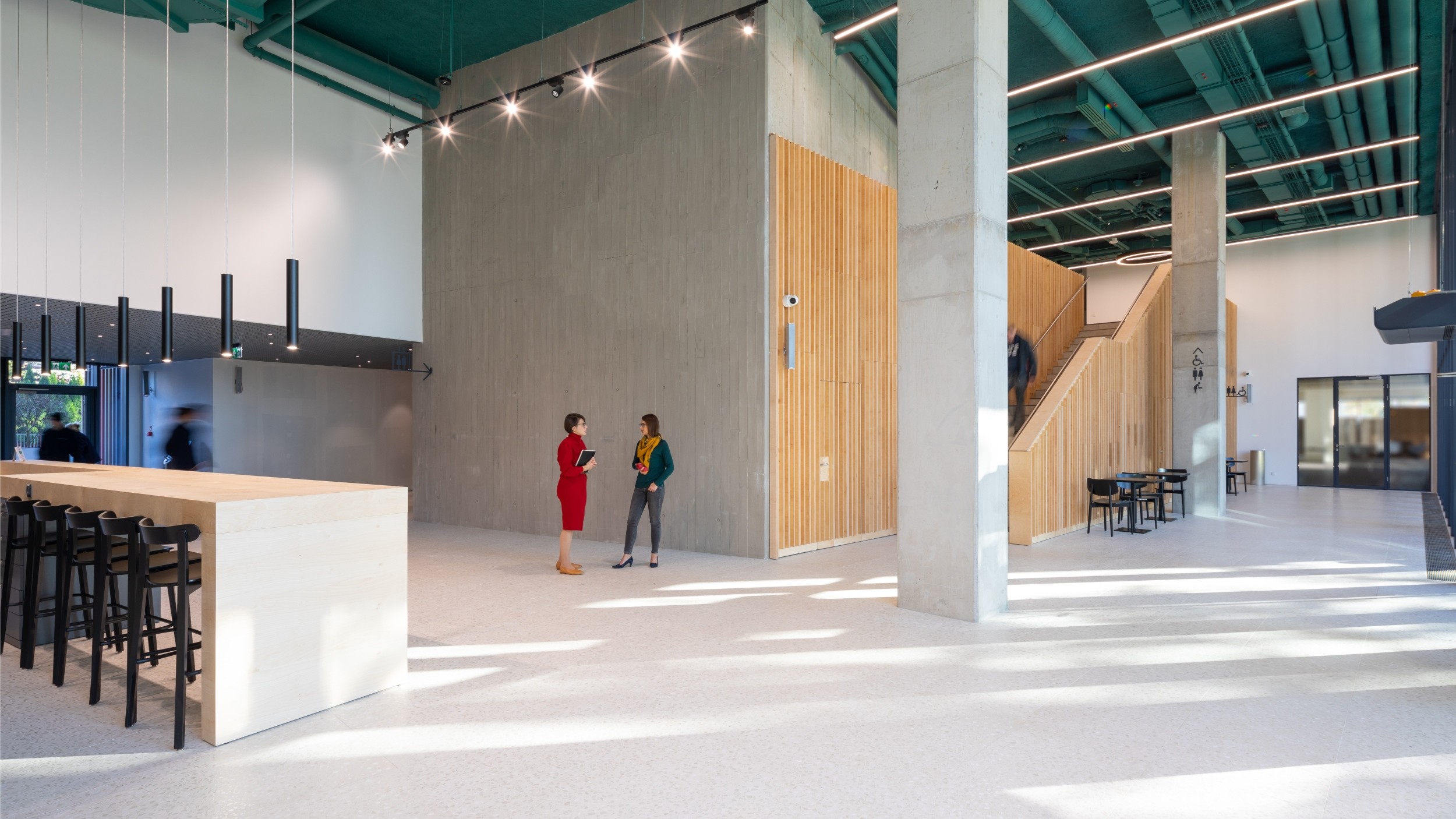 Modern lobby with high ceilings, wooden accents, and two people conversing near a staircase.