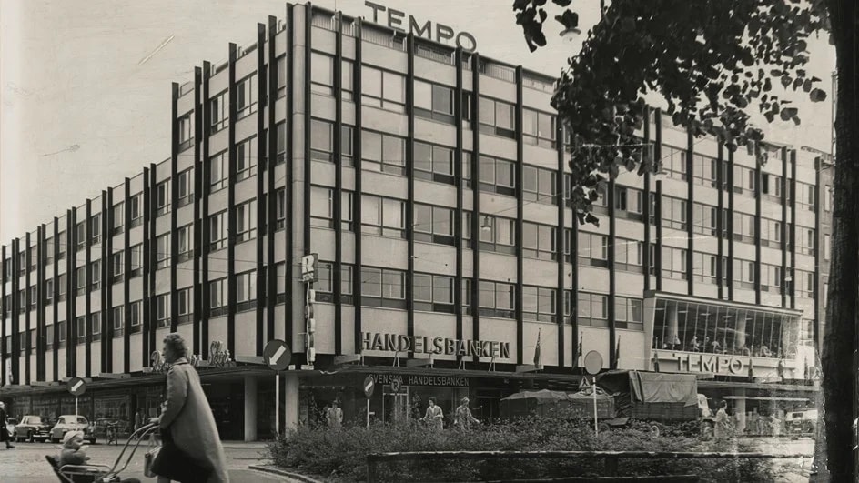 Svartvit bild på en kommersiell byggnad från mitten av århundradet med vintagebilar på gatan.