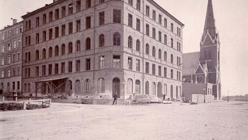 Historiskt svartvitt foto av en stor tegelbyggnad under byggnation bredvid en kyrka.
