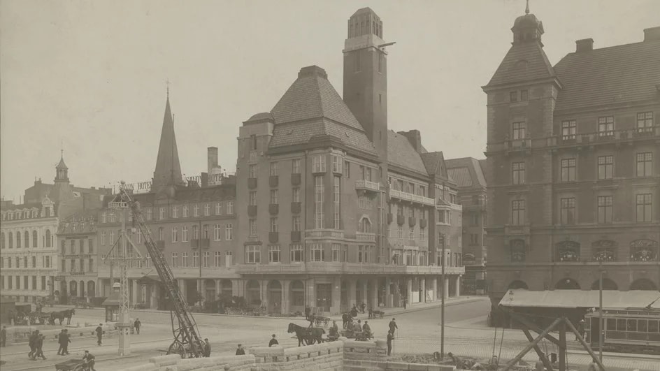Historiskt stads torg med vintage byggnader, hästdragna vagnar och tidigt 1900-talets spårvagn.