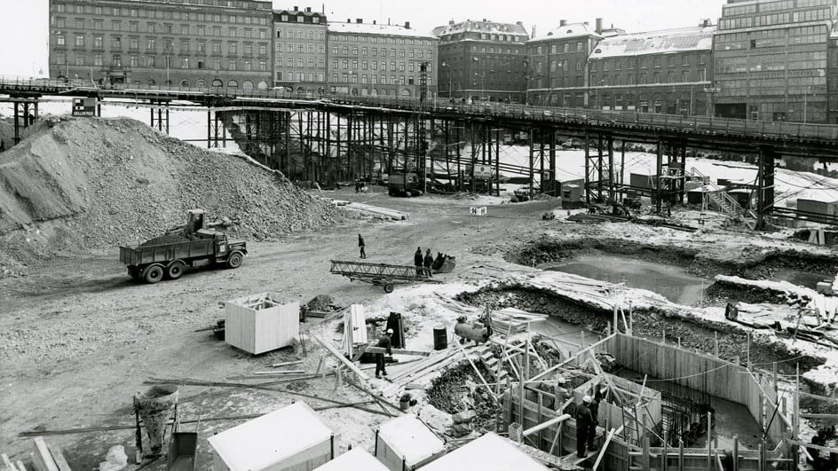 Byggarbetsplats med arbetare, utrustning och byggnader i bakgrunden.