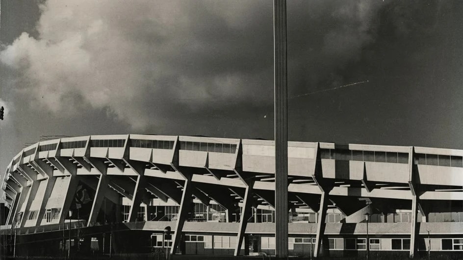 Svartvit bild av en stor, böjd stadionstruktur under en molnig himmel.