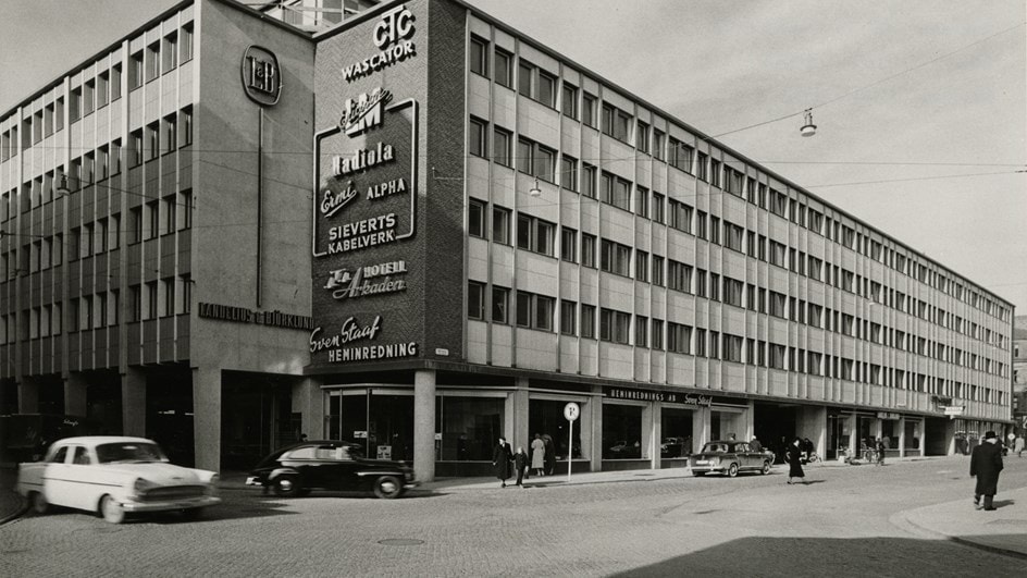 Svartvit foto av en kommersiell byggnad från mitten av århundradet med vintagebilar på gatan.