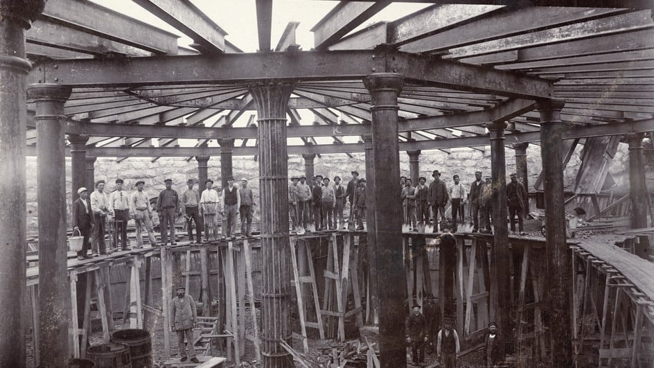 Historiskt svartvitt foto av arbetare som står inne på en stor industriell byggarbetsplats.