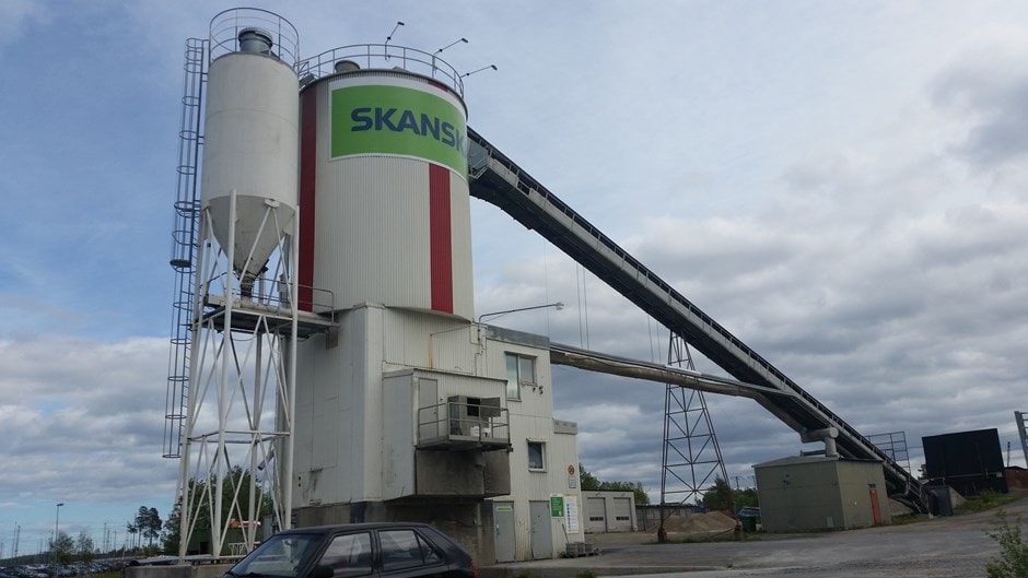Industrial cement silo and conveyor system at construction site Industriell cementsilo och transportband under en molnig himmel i en utomhusanläggning.