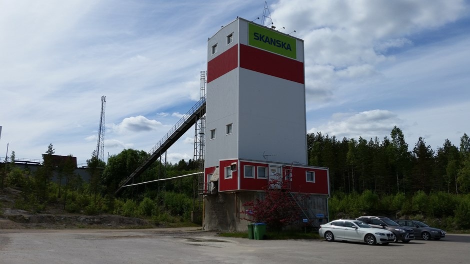 Skanska Industrial Building with Cars and Forest Background Hög industribyggnad med Skanska-logotyp, omgiven av träd och parkerade bilar.