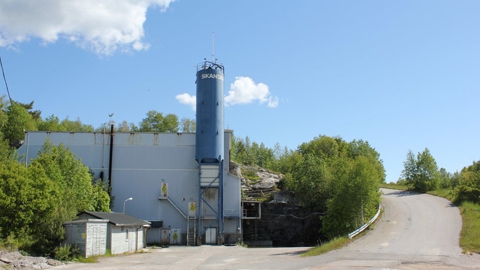 Industrial silo and storage facility near a rural road, clear sky Industriell silo och byggnad intill en slingrande väg omgiven av gröna träd och blå himmel.