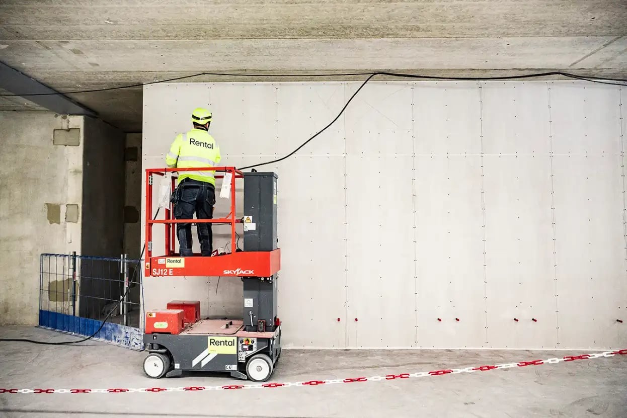 Arbetare i säkerhetsutrustning som manövrerar en röd saxlyft inuti en betongbyggnad under konstruktion.