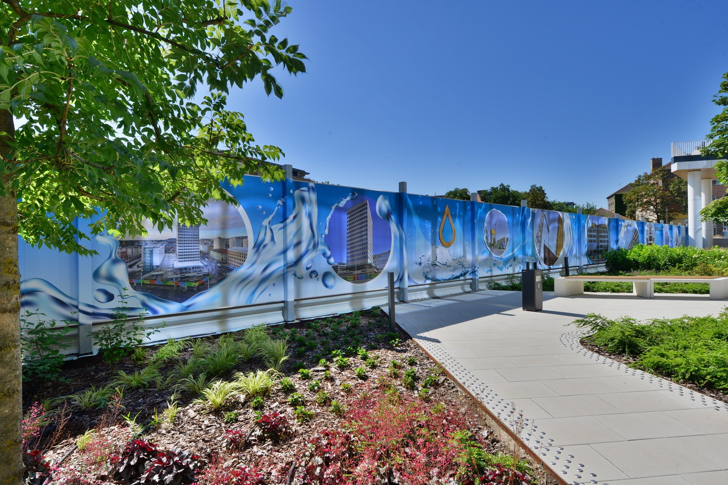 Colorful mural on a fence depicting water elements, surrounded by lush greenery and pathways.