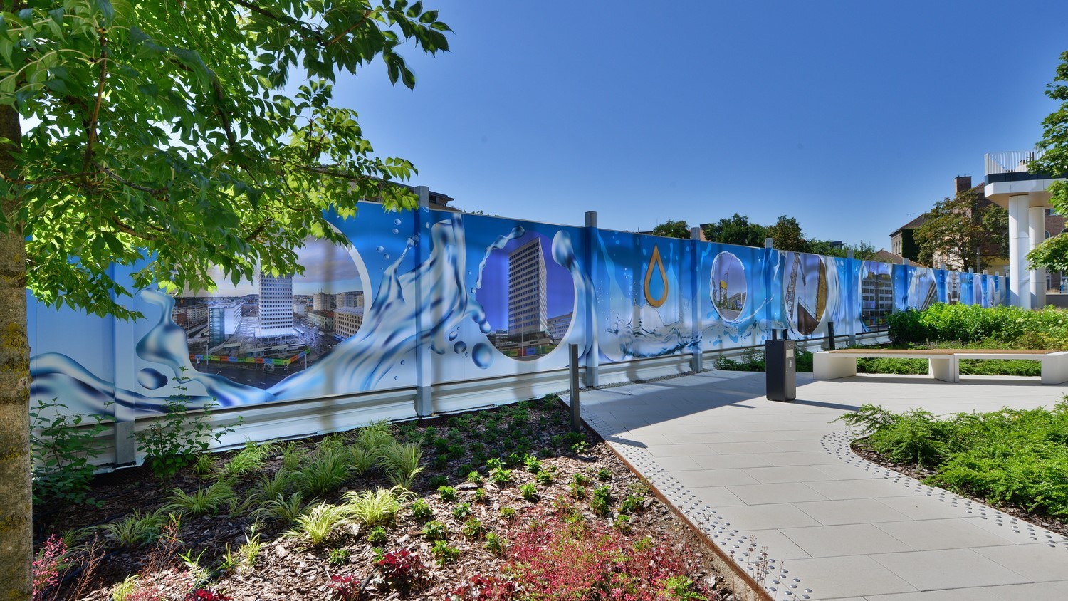 Colorful mural of water-themed images on a fence in a landscaped garden under a clear blue sky.