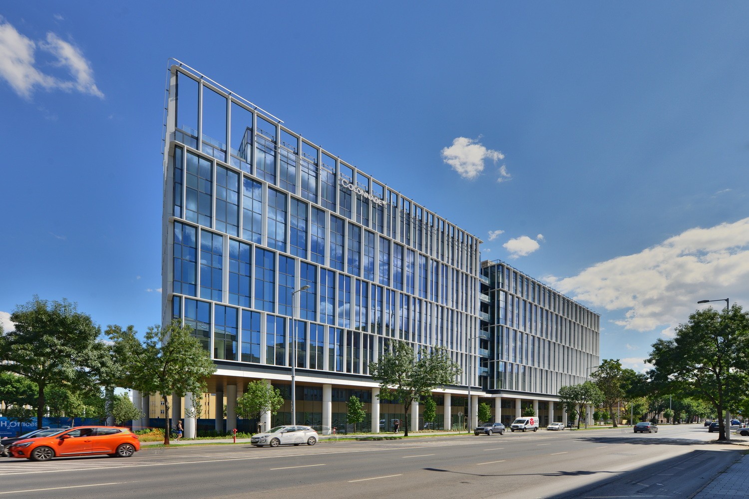 H₂Offices Phase 1 Modern glass office building with cars on a sunny street, under a clear blue sky.