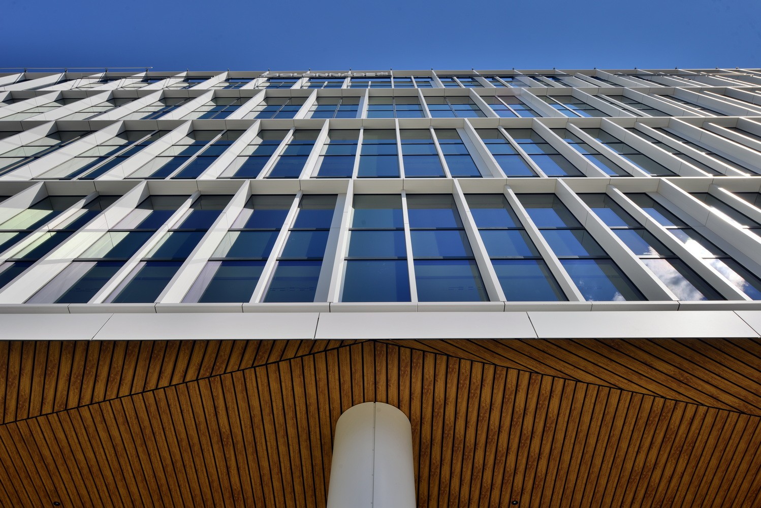 Modern building with glass facade and wood paneling, viewed from below against a clear blue sky.