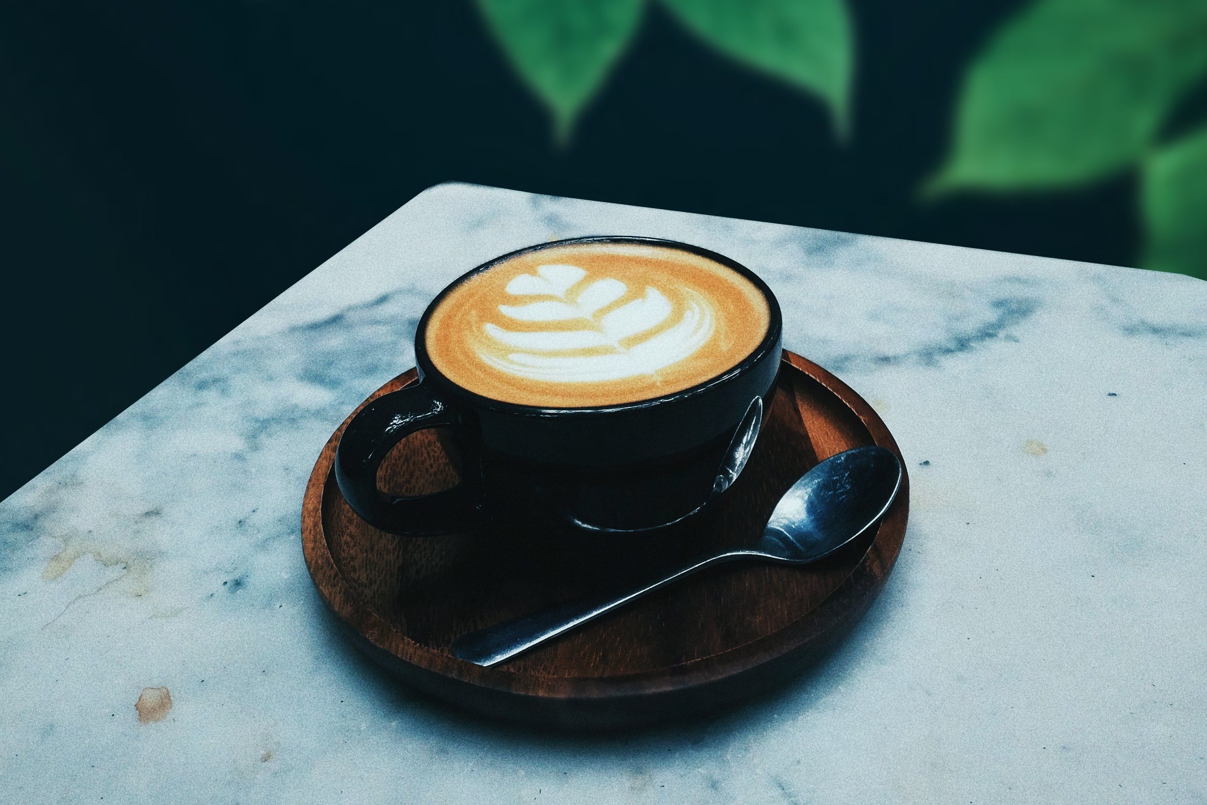 A cappuccino with latte art in a black cup on a wooden tray with a spoon, set on a marble table.