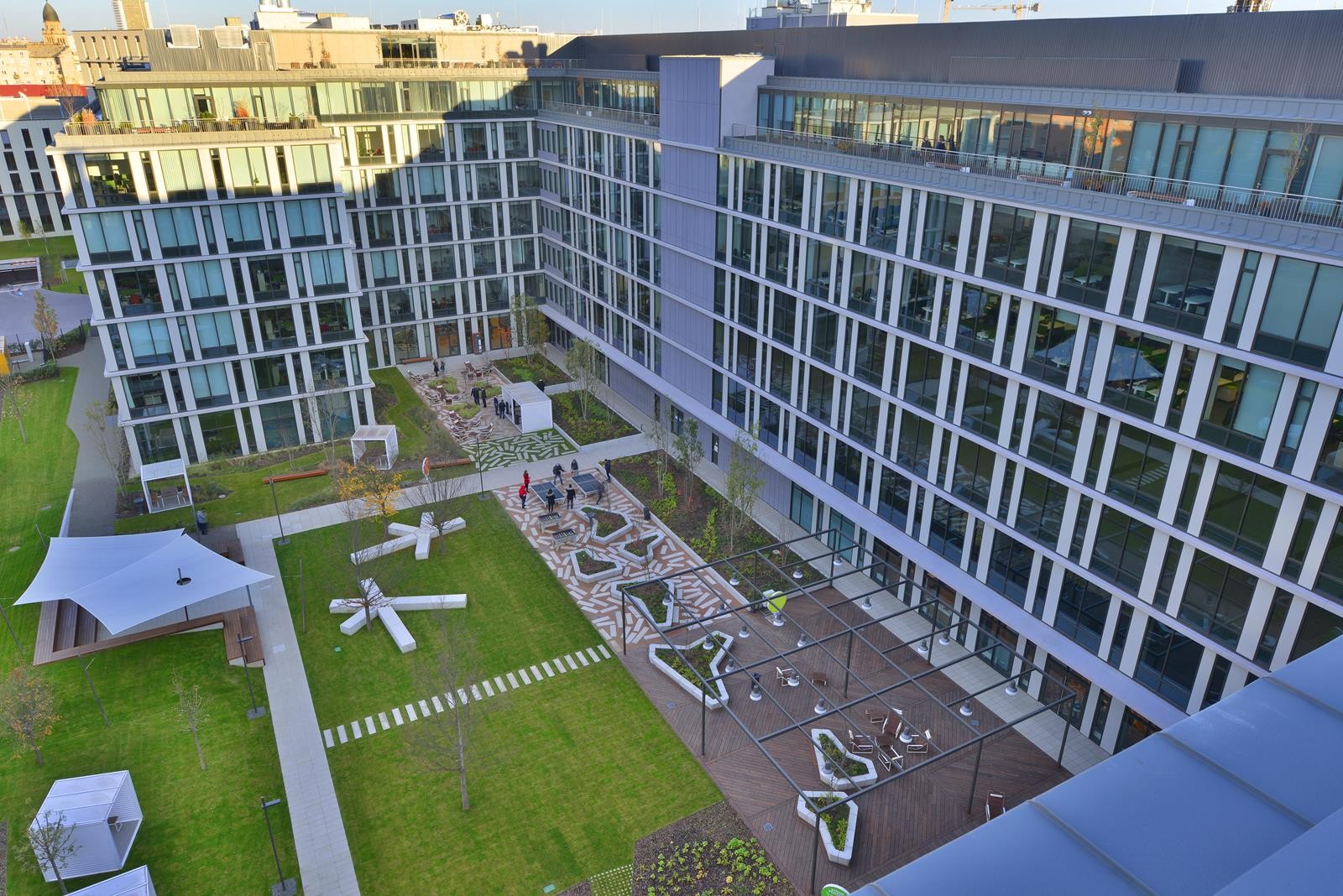 Modern urban courtyard with green spaces, seating areas, and surrounding glass-fronted buildings.
