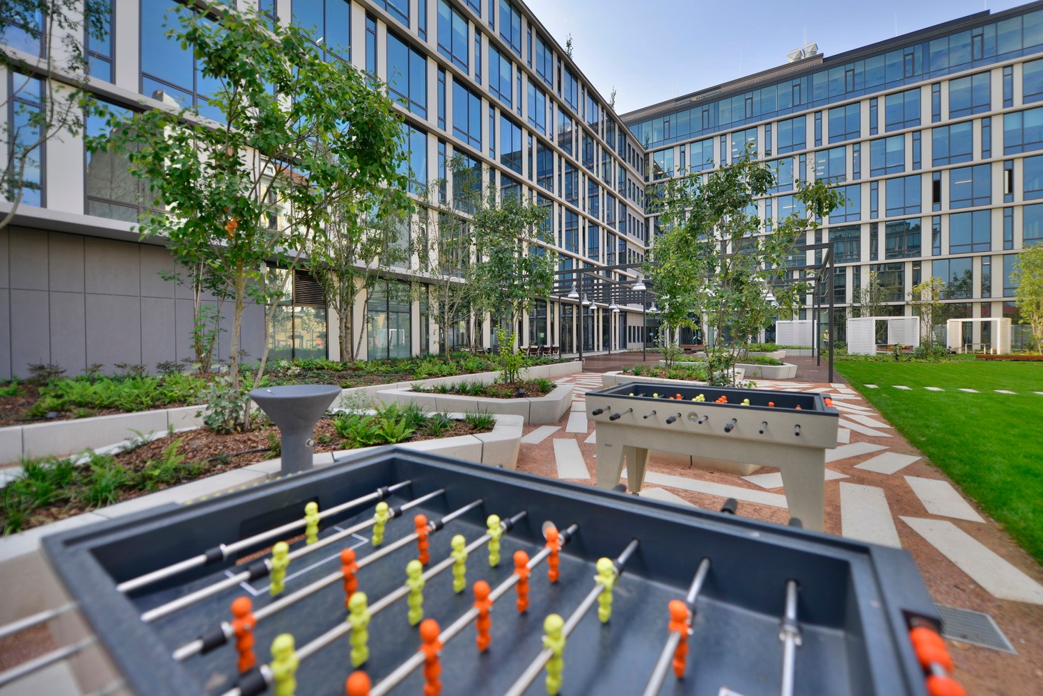 Modern office courtyard with foosball tables, greenery, and a glass building facade in the background.