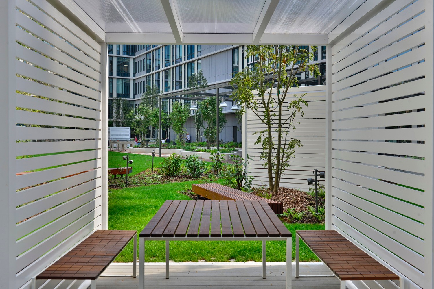 Modern outdoor seating area with wooden benches and table, surrounded by lush greenery and buildings.