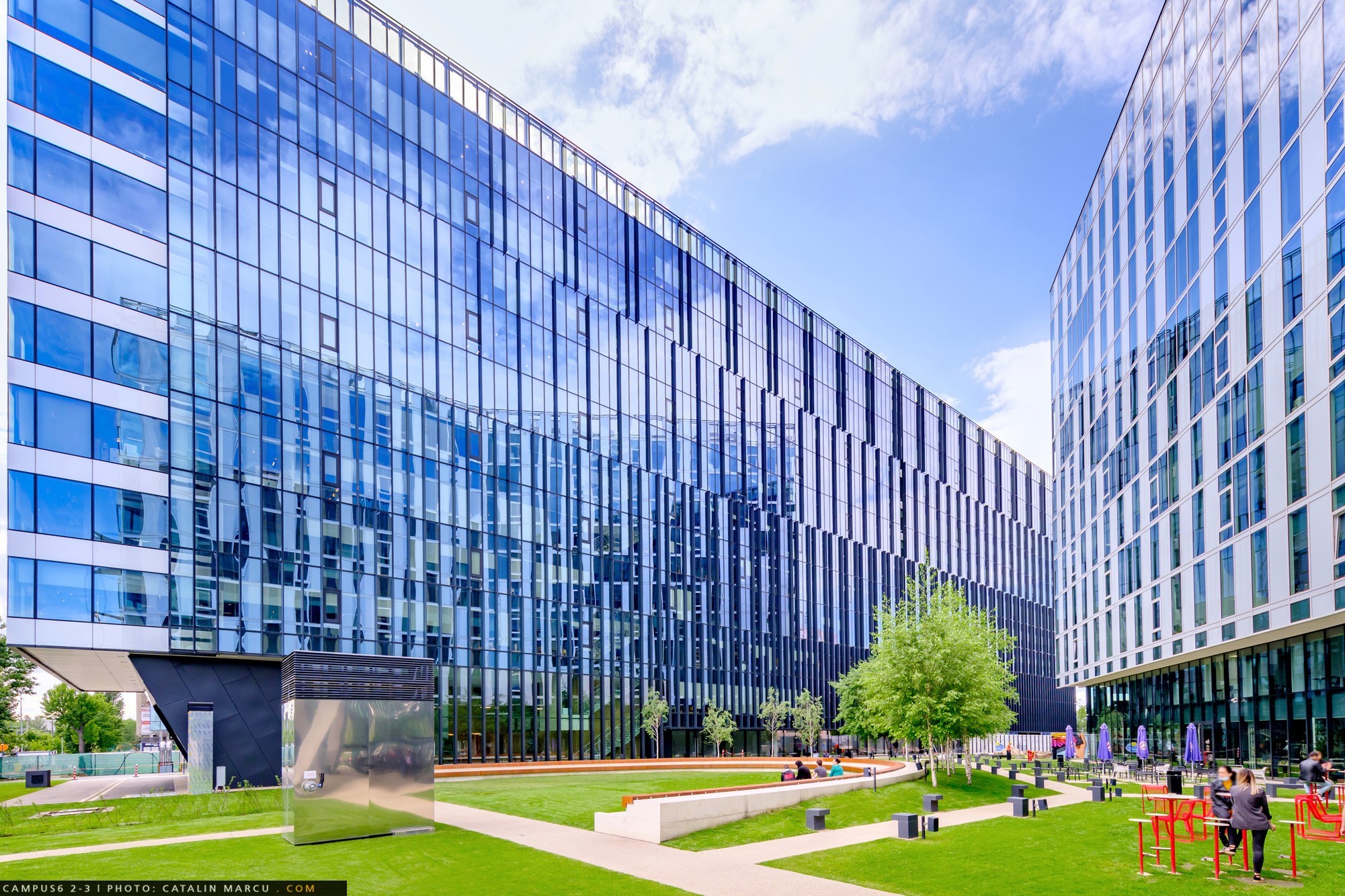 Campus 6 Modern glass buildings surrounding a landscaped courtyard with trees and seating in sunny weather.