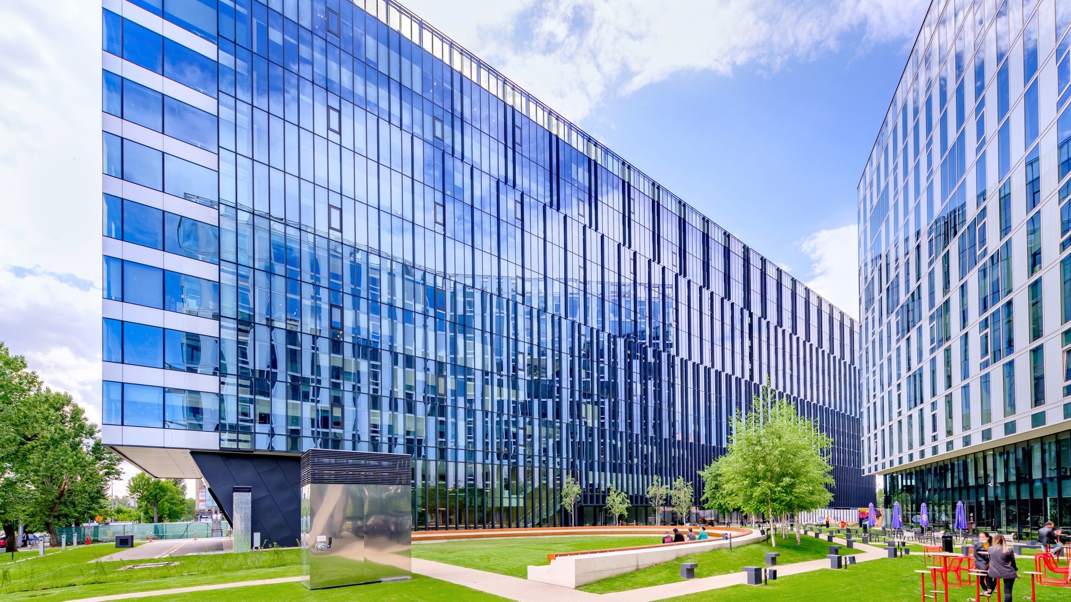 Campus 6.2&6.3 Modern glass office buildings with green lawn and outdoor seating, under a partly cloudy sky.