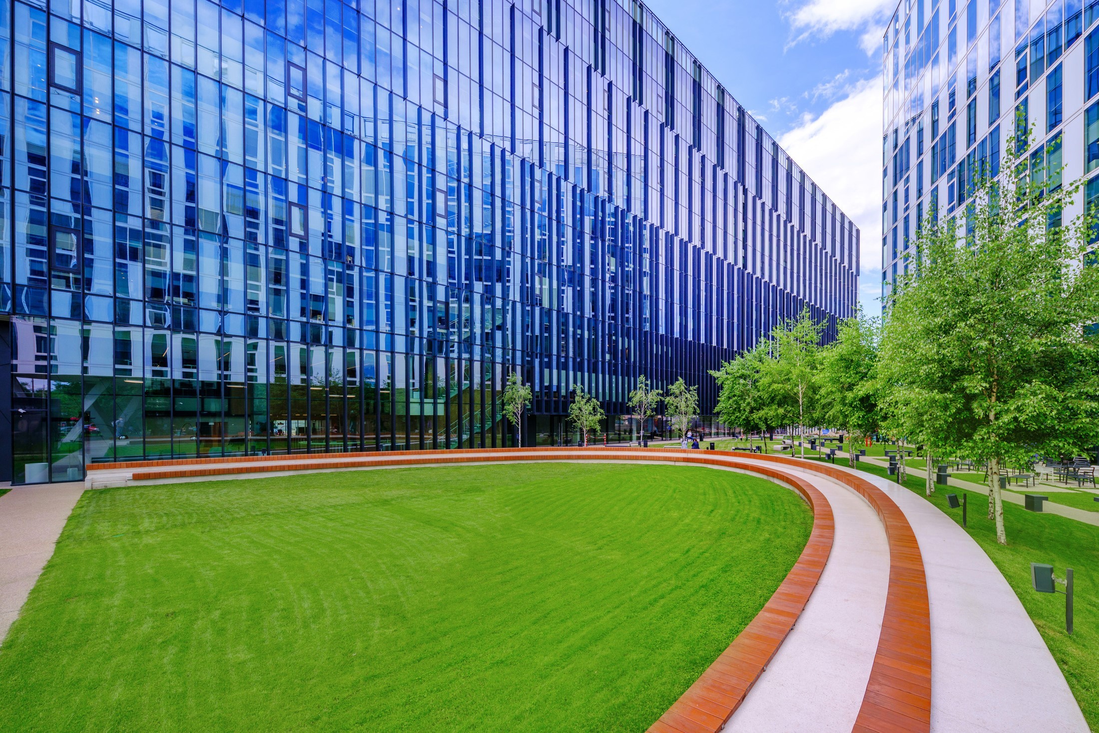 Modern glass building with a curved lawn and benches, surrounded by trees and a pathway.