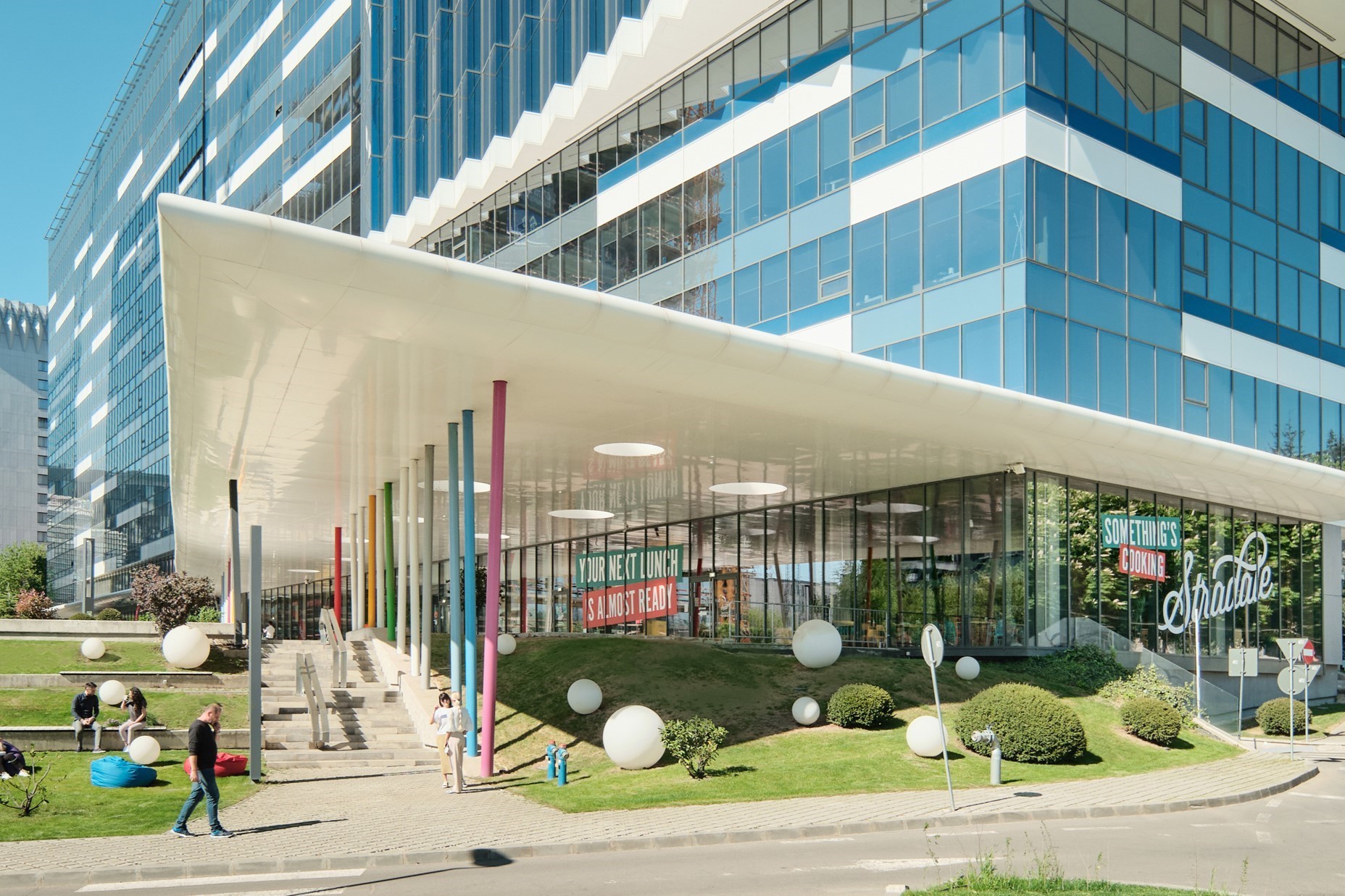 Equilibrium Modern glass building with colorful pillars, landscaped lawn, and people walking outside.