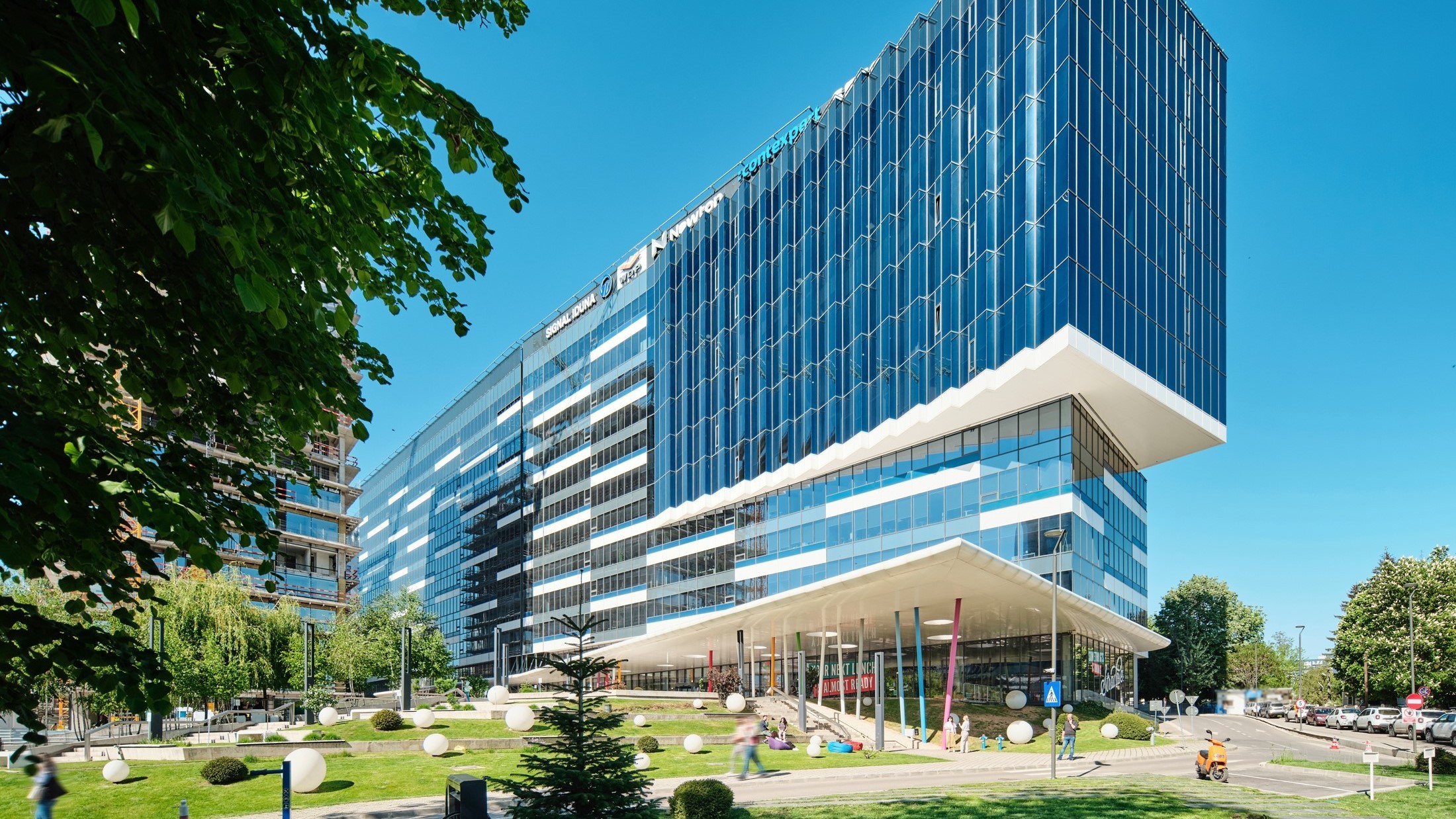 Modern glass office building with a unique angular design surrounded by trees and a landscaped area.
