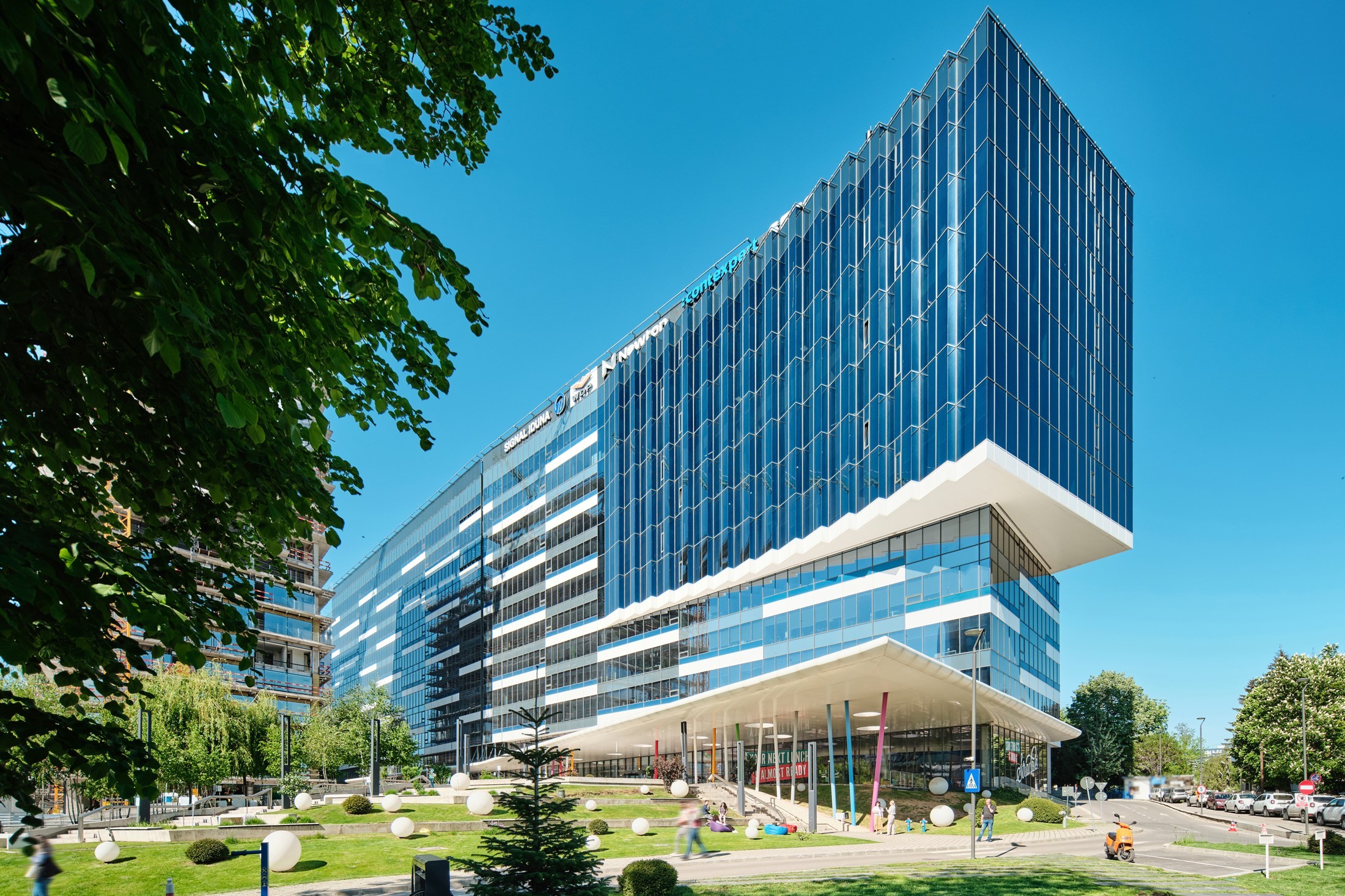 Equilibrium Modern glass office building with a unique overhang design, surrounded by greenery and pathways.