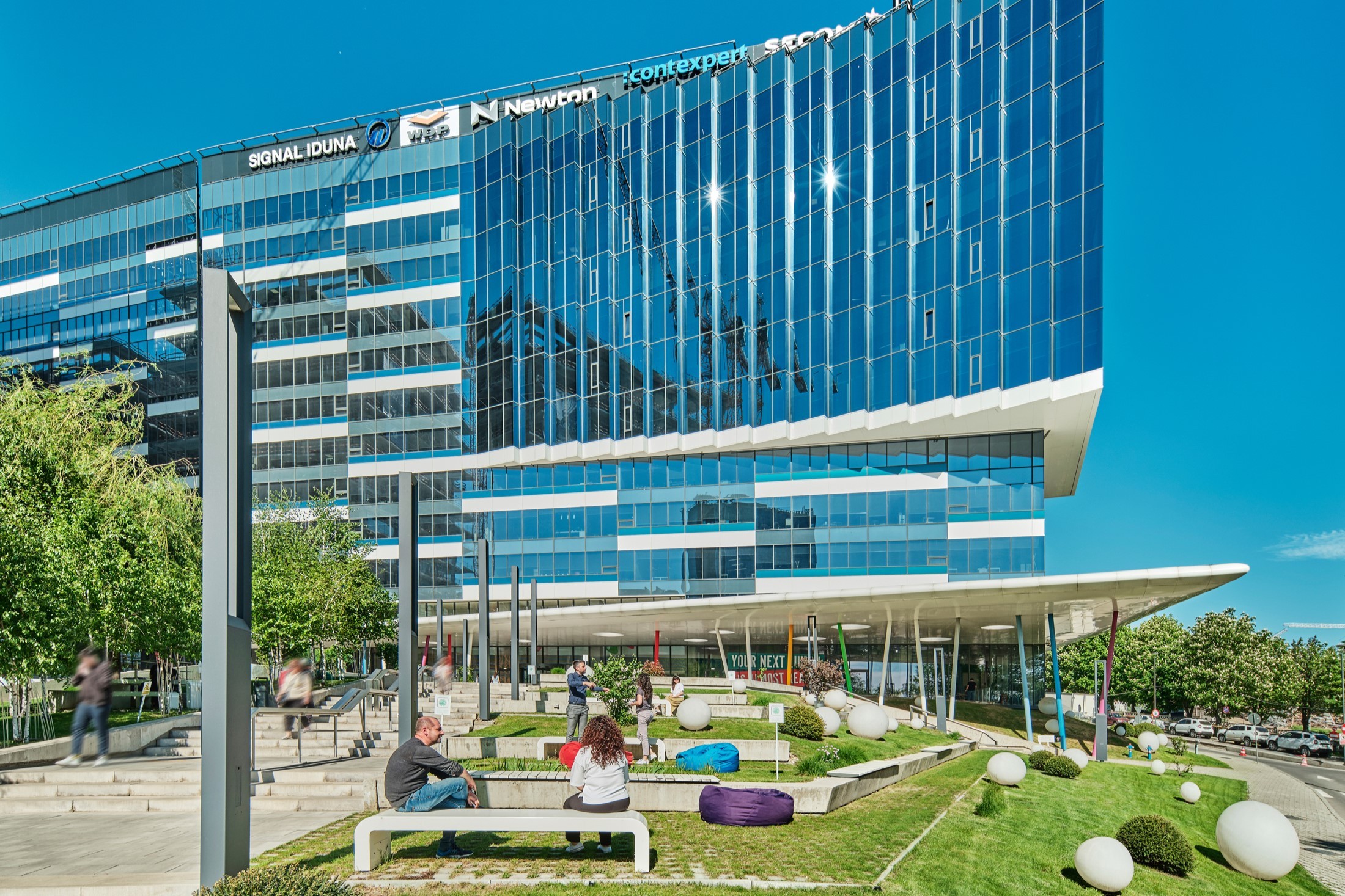 Modern glass office building with landscaped outdoor seating area, featuring lush greenery.