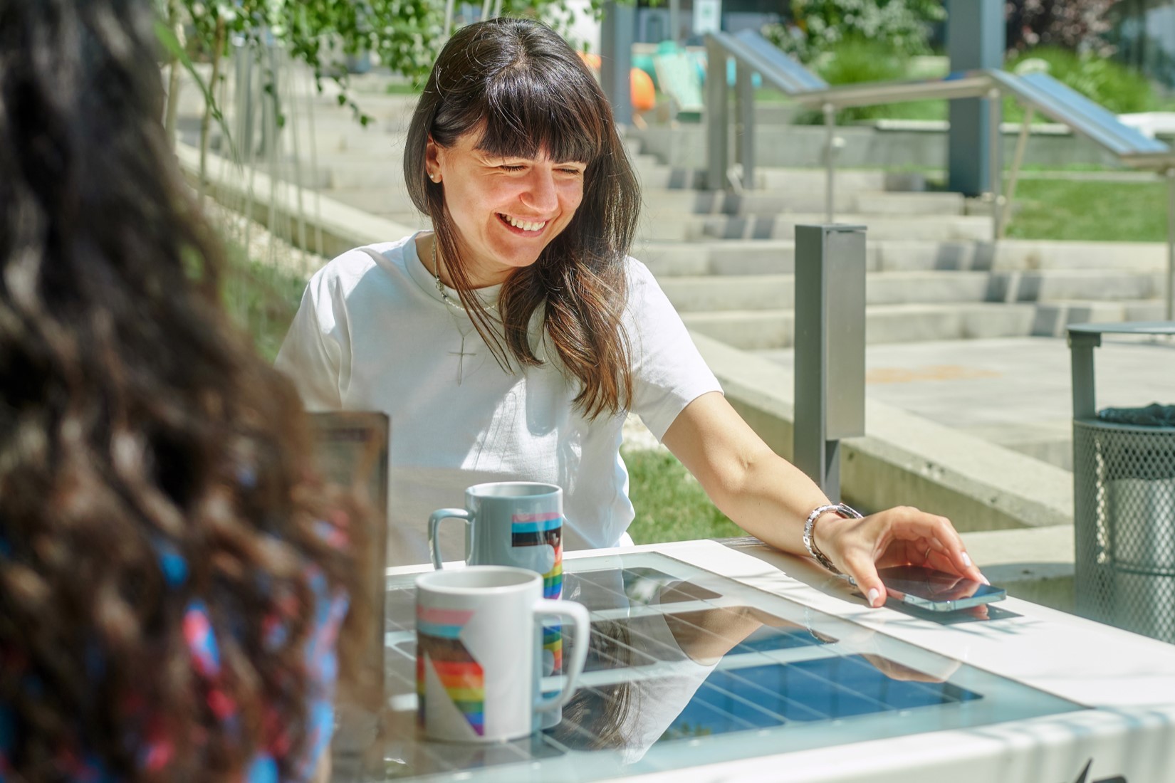 Two individuals sit at a sunny outdoor table with rainbow mugs and a smartphone, surrounded by greenery.