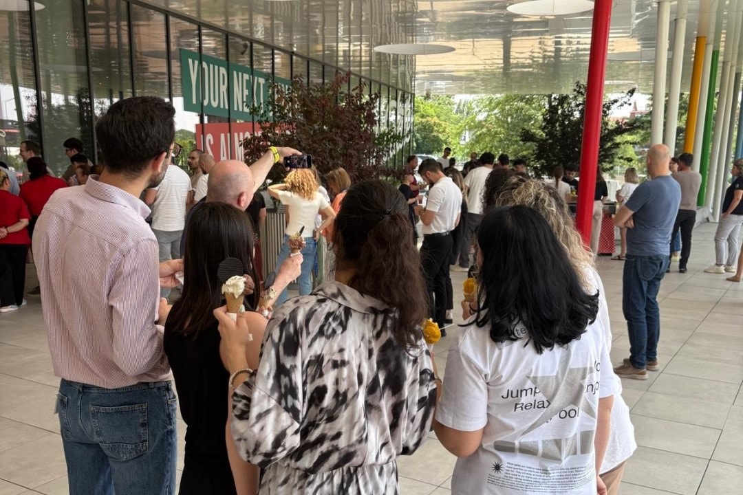A group of people waits in line outdoors at a mobile coffee cart near a glass building.