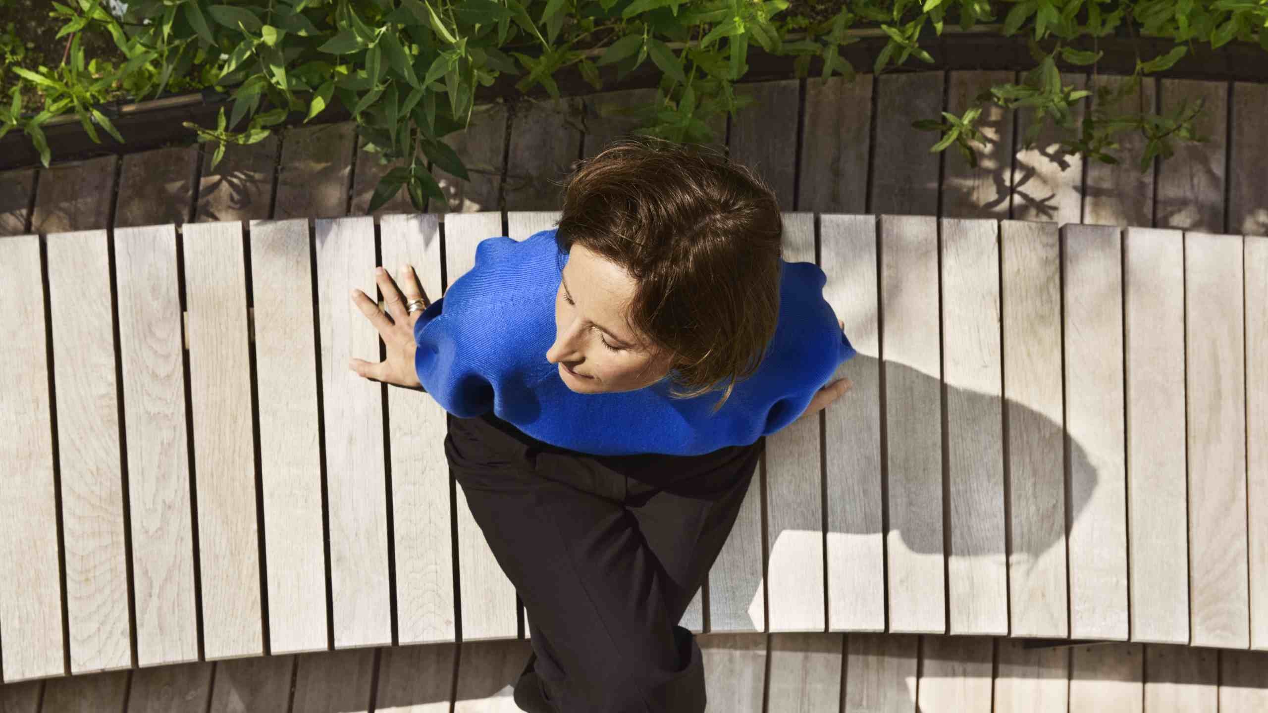 A person wearing a blue top sits on a curved wooden bench surrounded by greenery.