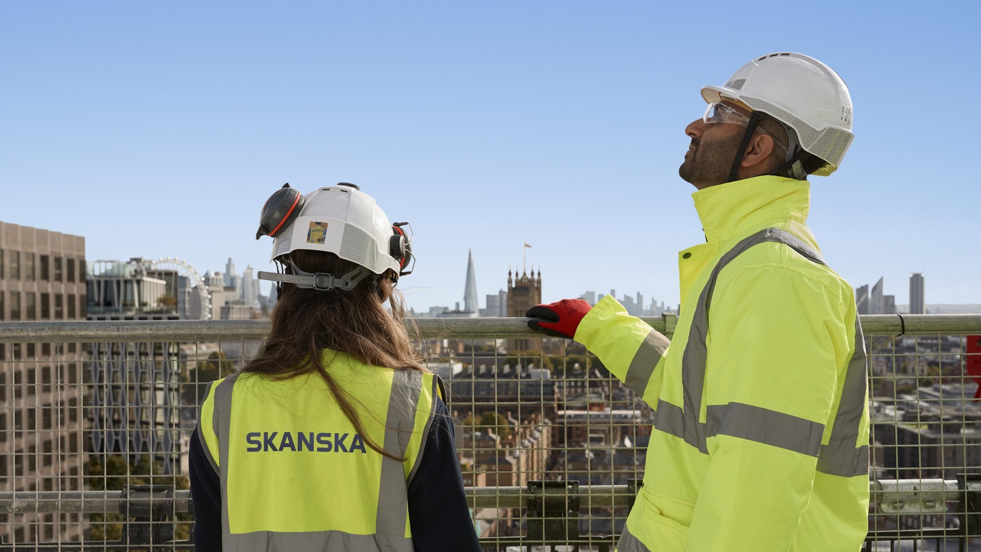 Construction Workers in Safety Gear at Skanska Job Site Construction workers in yellow safety gear and helmets converse at a Skanska site.