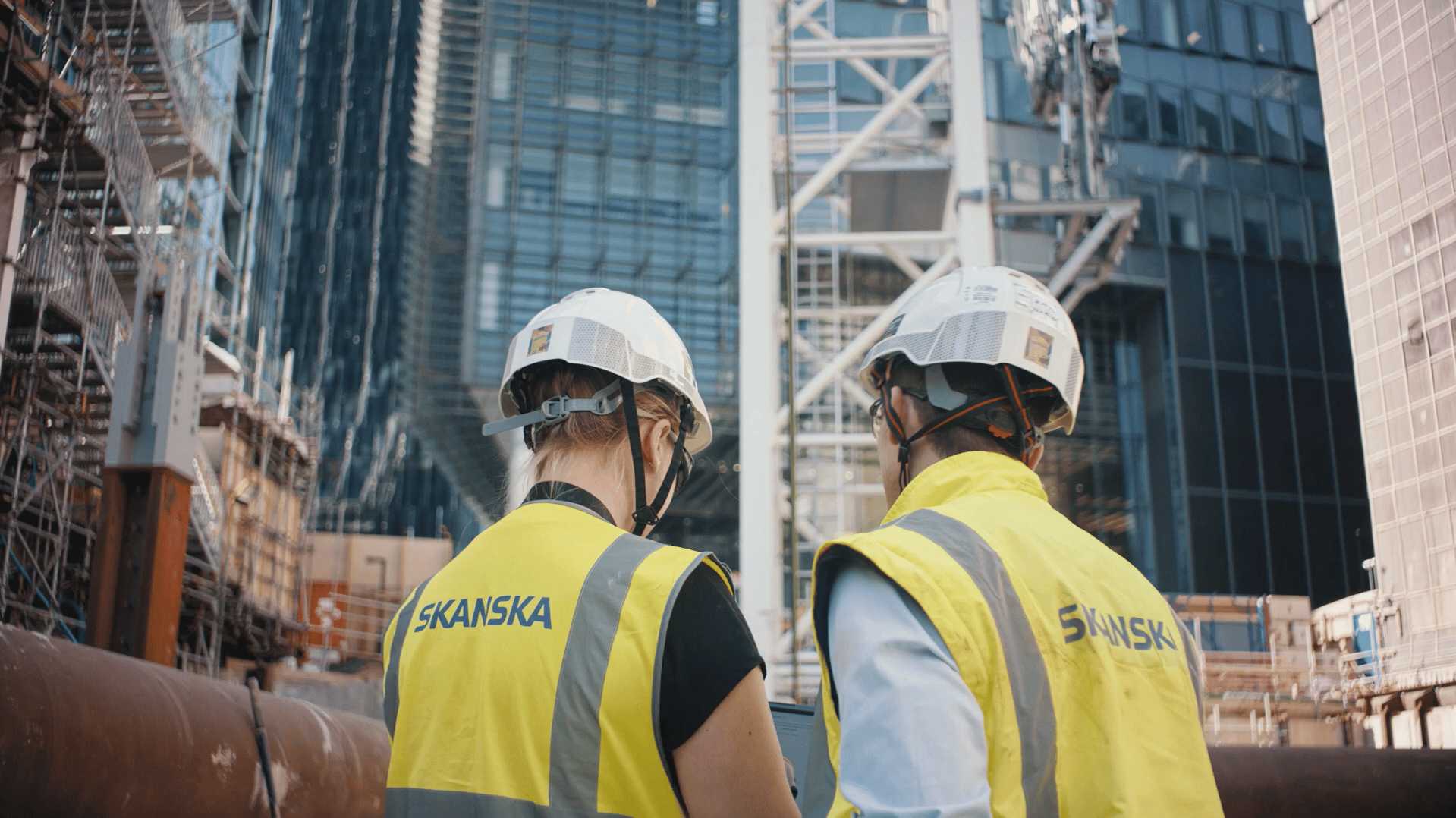 Two construction workers wearing Skanska vests and helmets at an urban building site.
