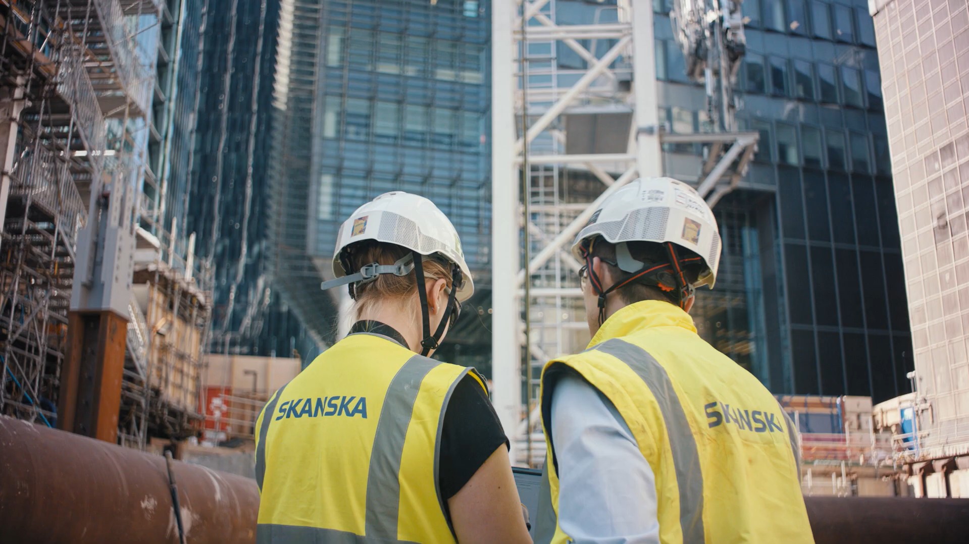 Two construction workers wearing Skanska vests and helmets at an urban building site.