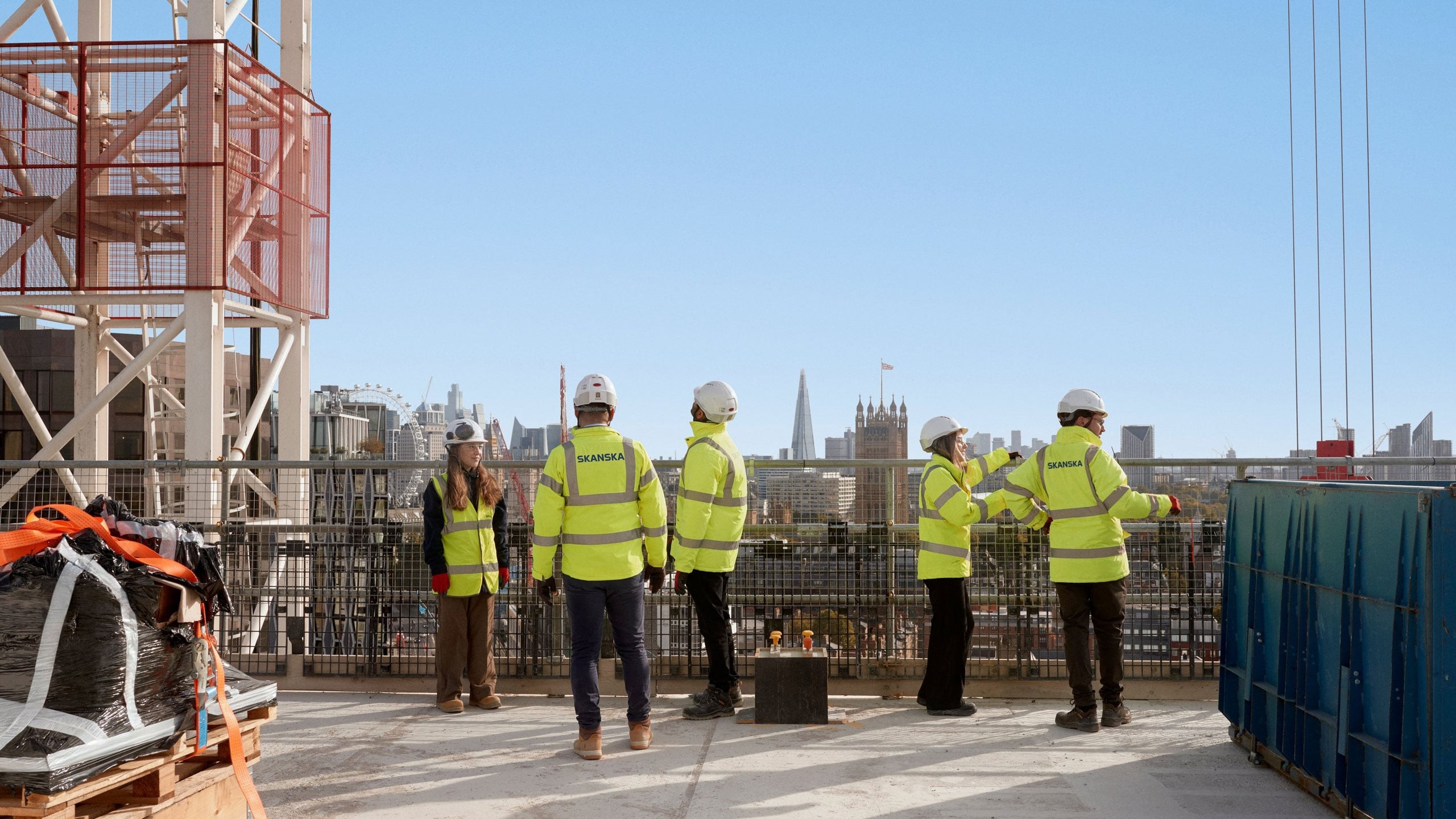Construction workers in high-visibility gear stand on a rooftop, overlooking a city skyline.