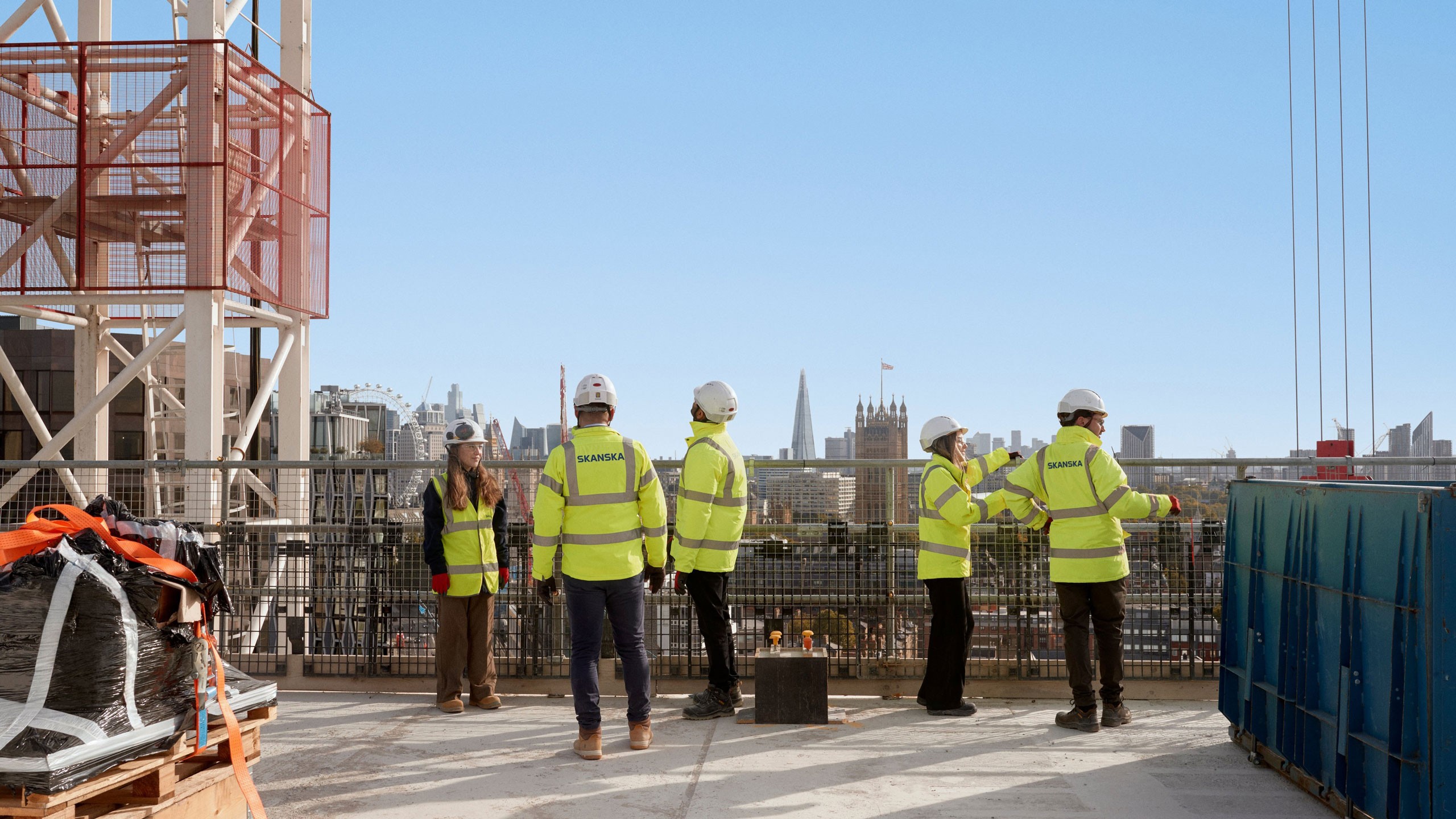 Construction workers in high-visibility gear stand on a rooftop, overlooking a city skyline.