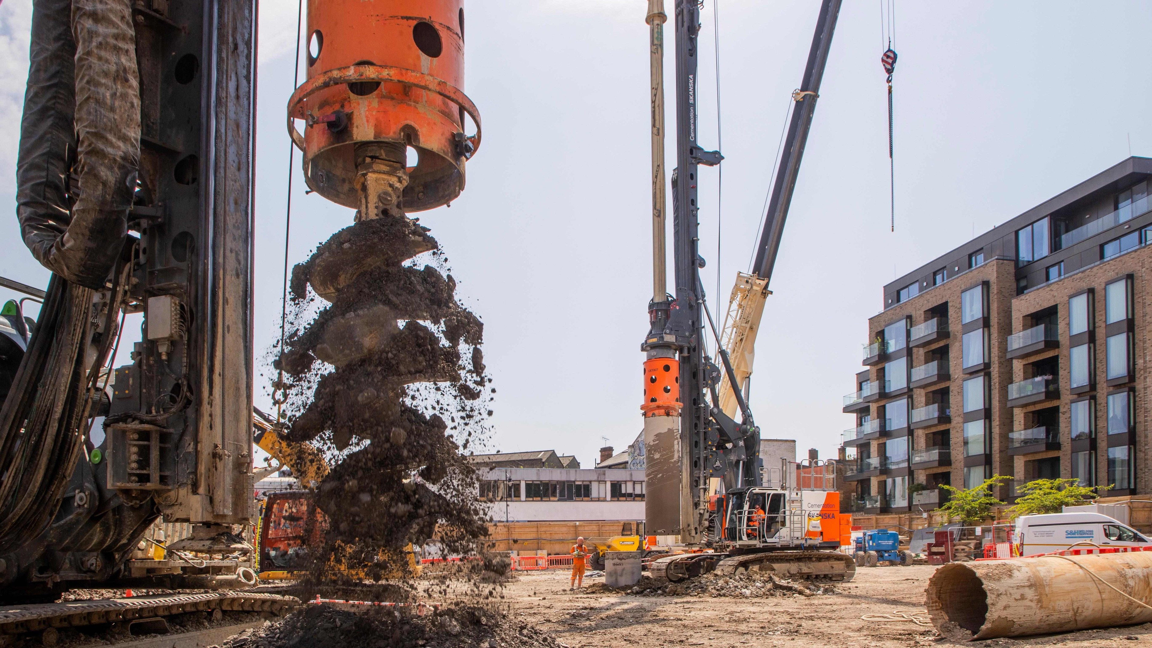 Construction site with heavy machinery drilling soil, modern buildings in the background.