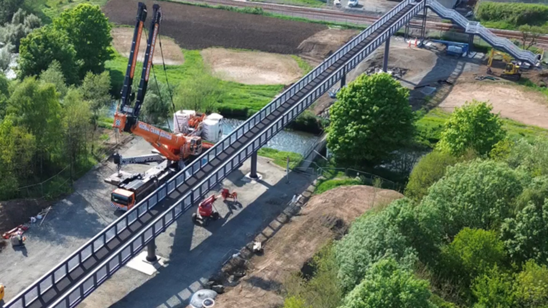 Construction of Elevated Bridge with Heavy Machinery Aerial view of a construction site with a long pedestrian bridge, cranes, and greenery surrounding.