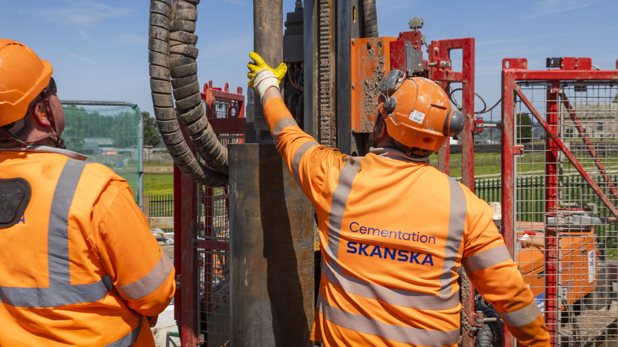 Construction workers operating cementing equipment on site Two construction workers in orange safety gear operate heavy machinery on a sunny day.