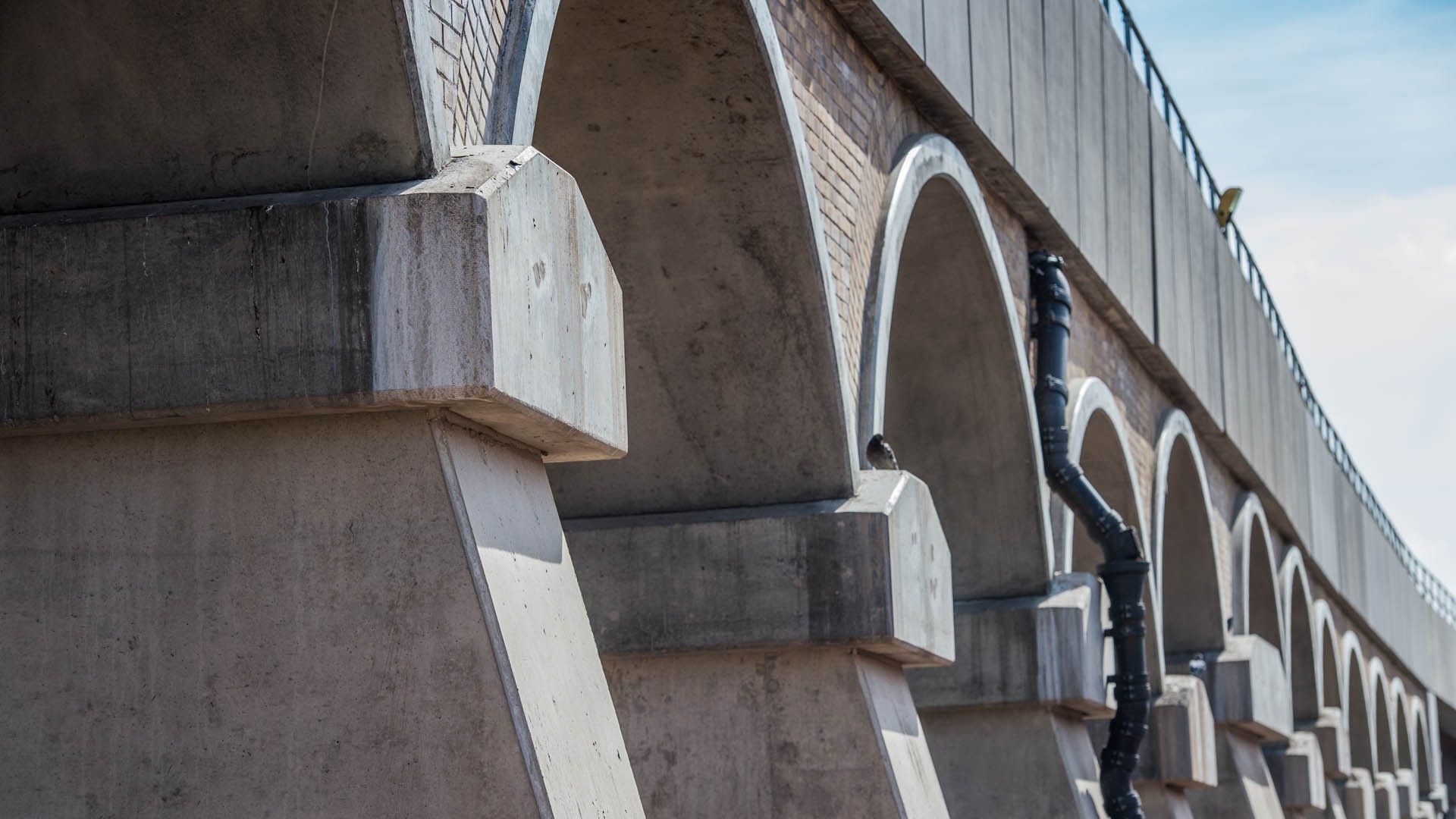 Concrete arched bridge with visible piping and textured brick detailing under a clear blue sky.