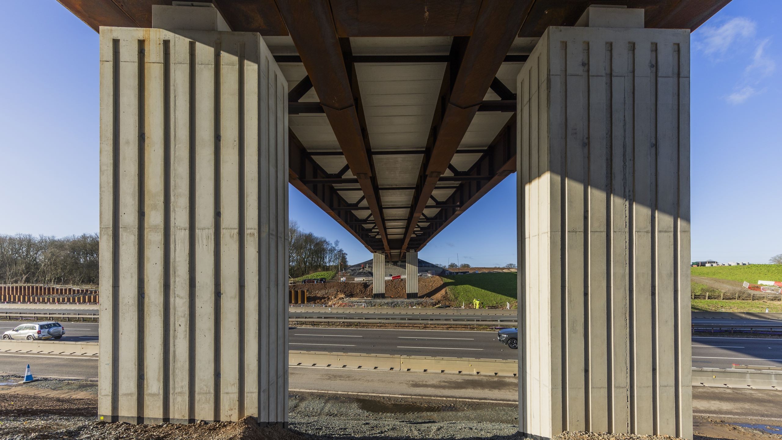 M42 J6 bridge View beneath a concrete highway bridge showing pillars, road, and clear blue sky.
