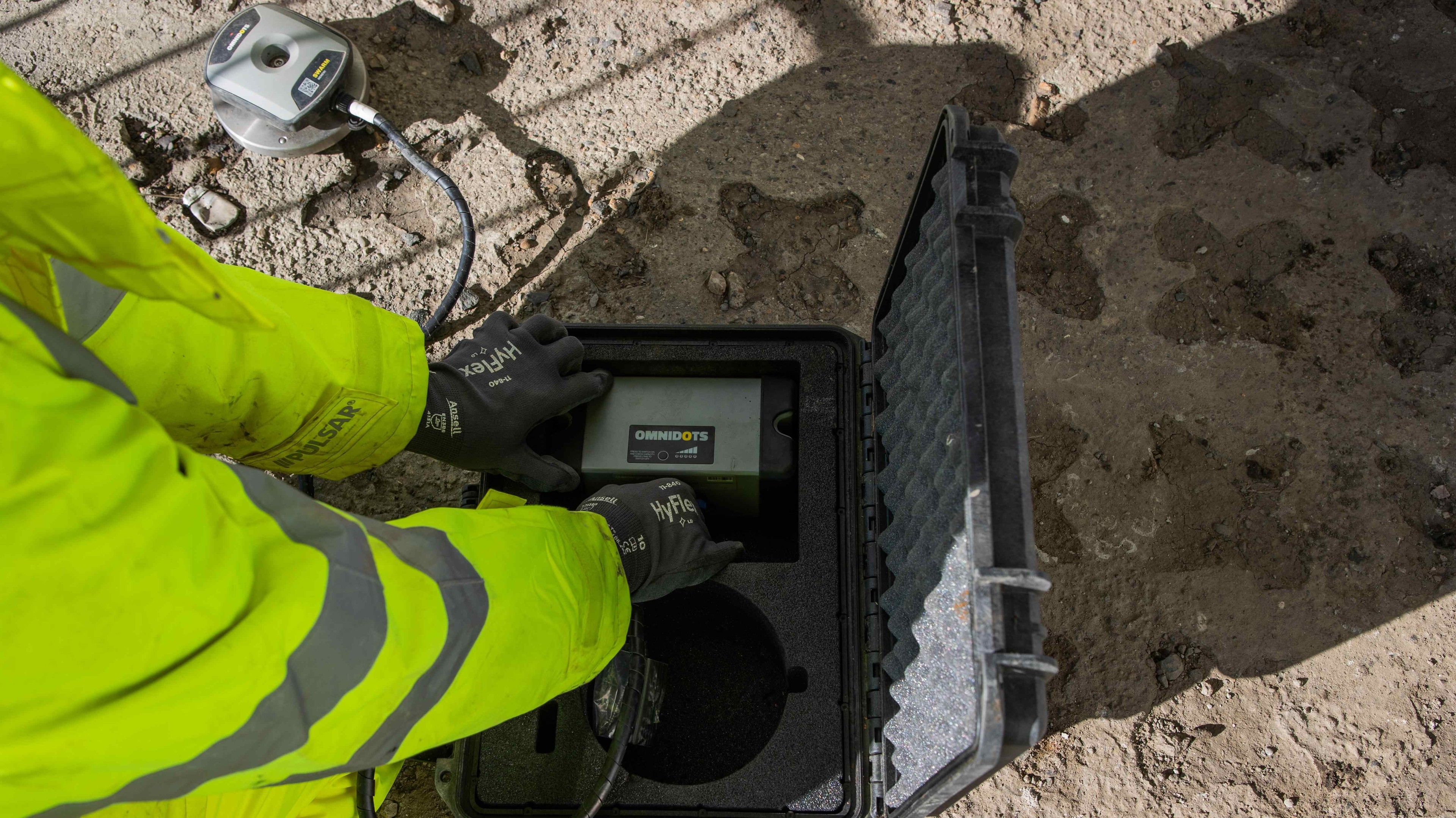 Construction Site Equipment: Worker Using Monitoring Device A person in a neon jacket uses a portable device from a rugged case on a dirt surface.