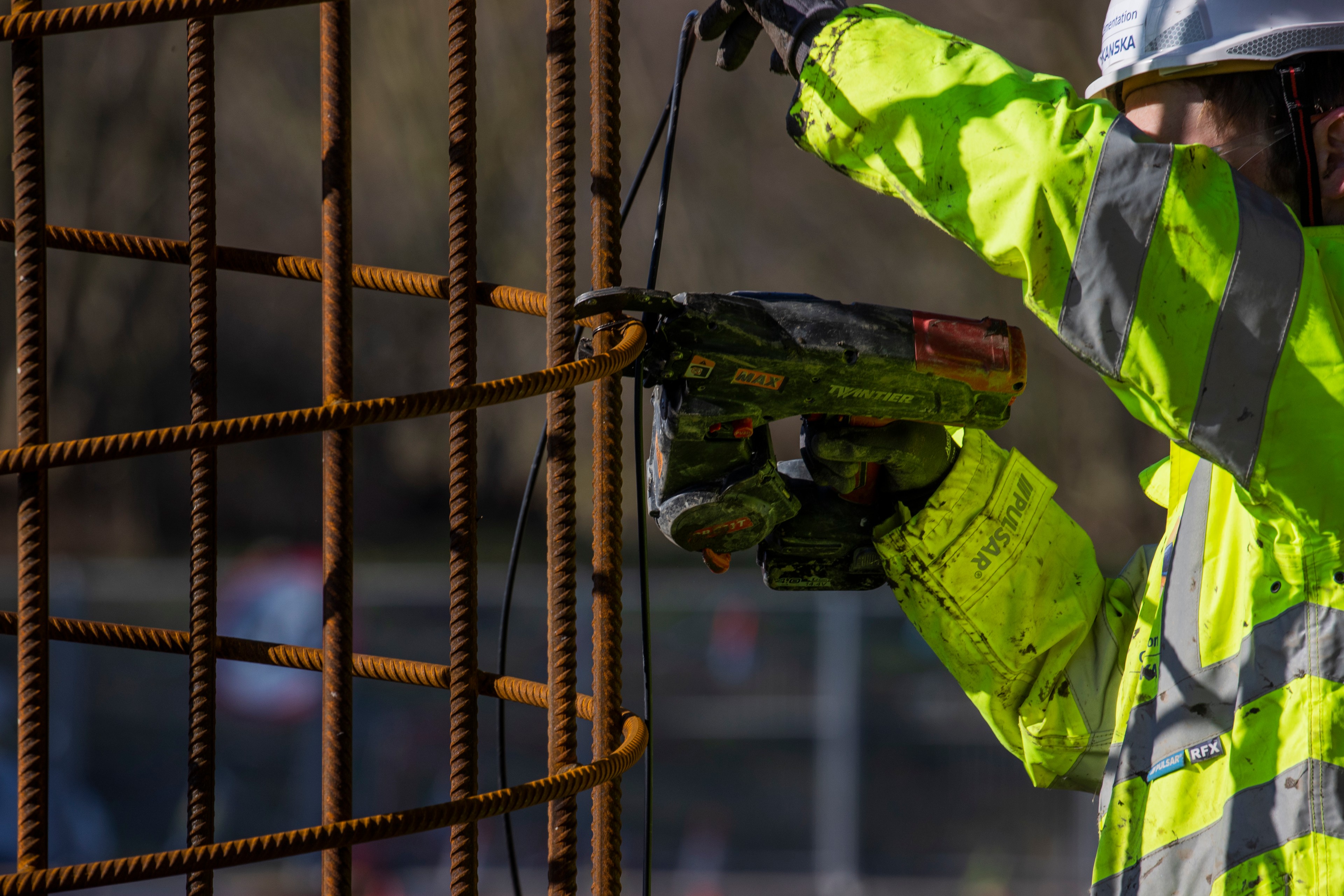 Construction Worker Securing Rebar with Power Tool Construction worker in high-visibility gear using a power tool on steel rebar structure outdoors.