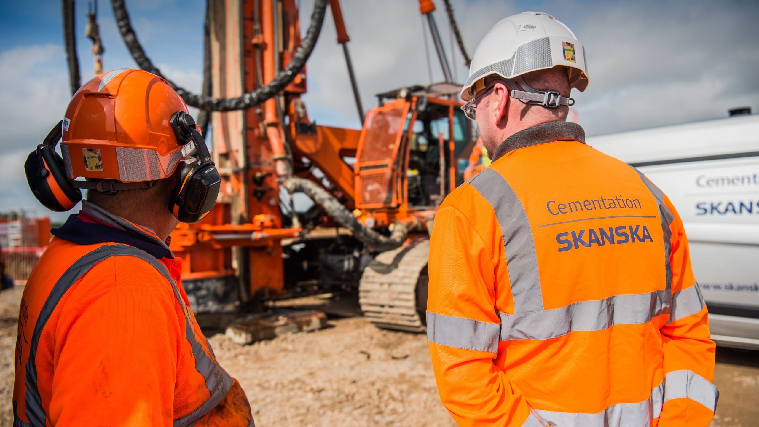 Construction workers in safety gear at Skanska site Two construction workers in safety gear overseeing heavy machinery at a building site.