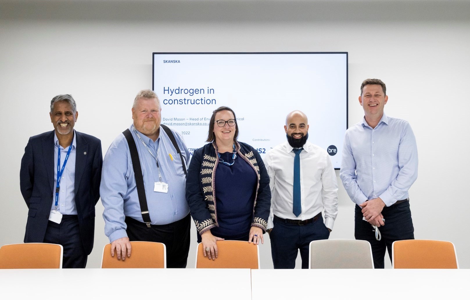 Team discussing hydrogen in construction presentation meeting Five professionals stand in a meeting room with a screen showing "Hydrogen in construction."