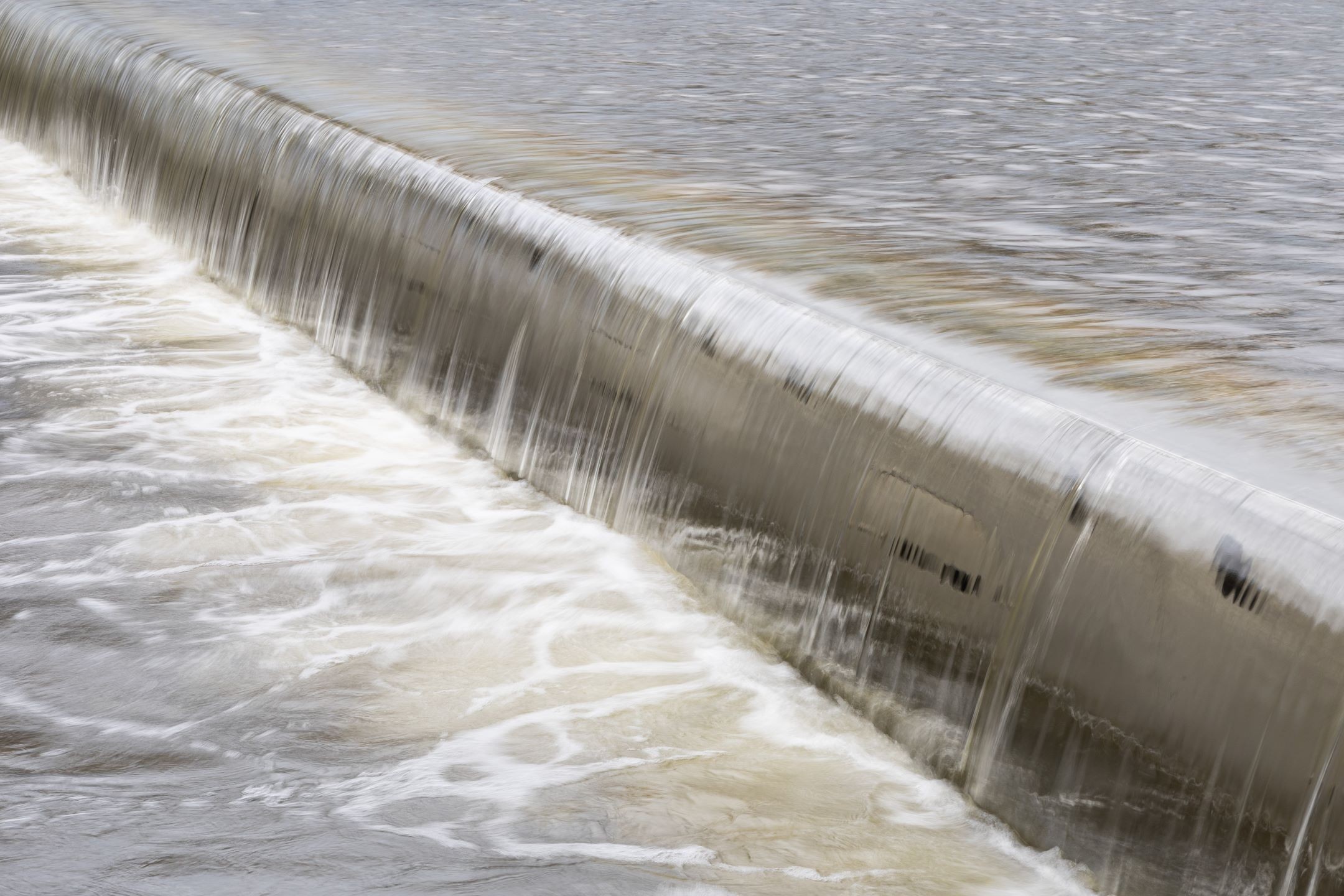 Smooth Flowing Water Over Concrete Weir in River Stream Overflowing water cascading smoothly over a concrete dam structure in a river.