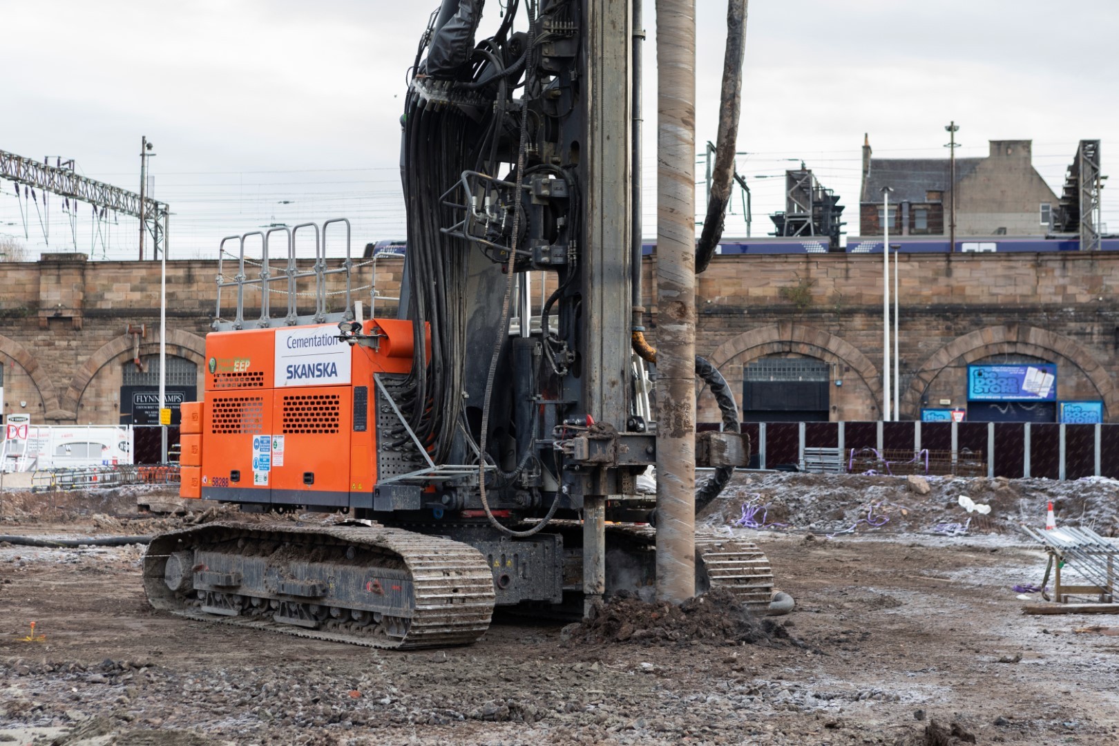 Construction site with a Skanska drilling rig on muddy ground, near an urban railway backdrop.