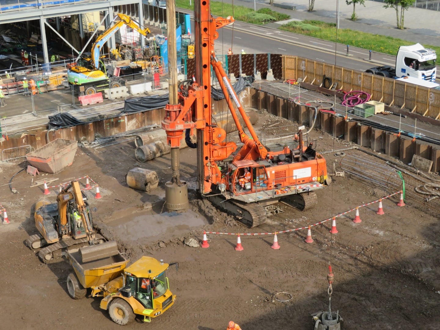 Construction site with heavy machinery, including an orange drilling rig and a yellow dumper truck.