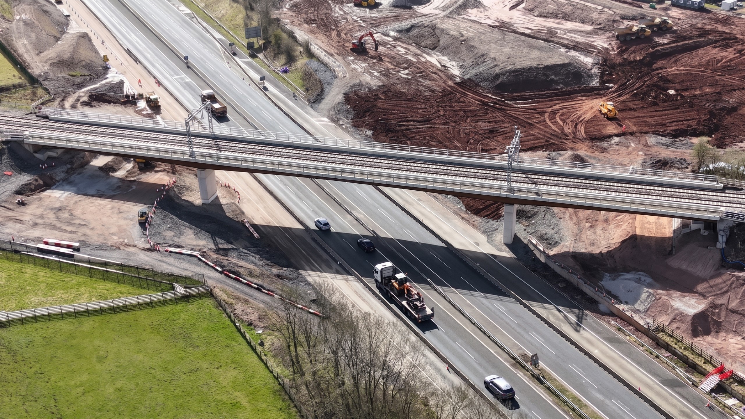 Modern Construction Site Amidst Scenic Countryside Views Aerial view of a highway with vehicles passing under a railway bridge and construction site.