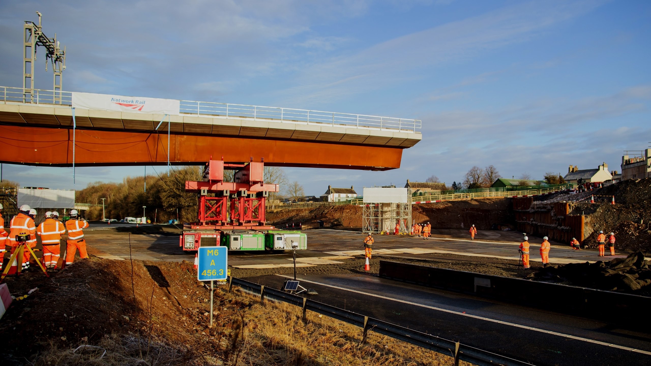 Construction workers install a large bridge segment over a highway at a roadside site.